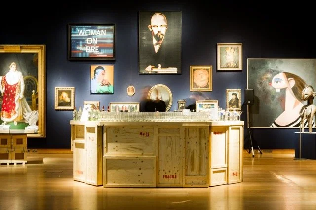 Art gallery interior with various paintings and sculptures displayed on dark walls, featuring portraits and abstract art, with a wooden central display table.
