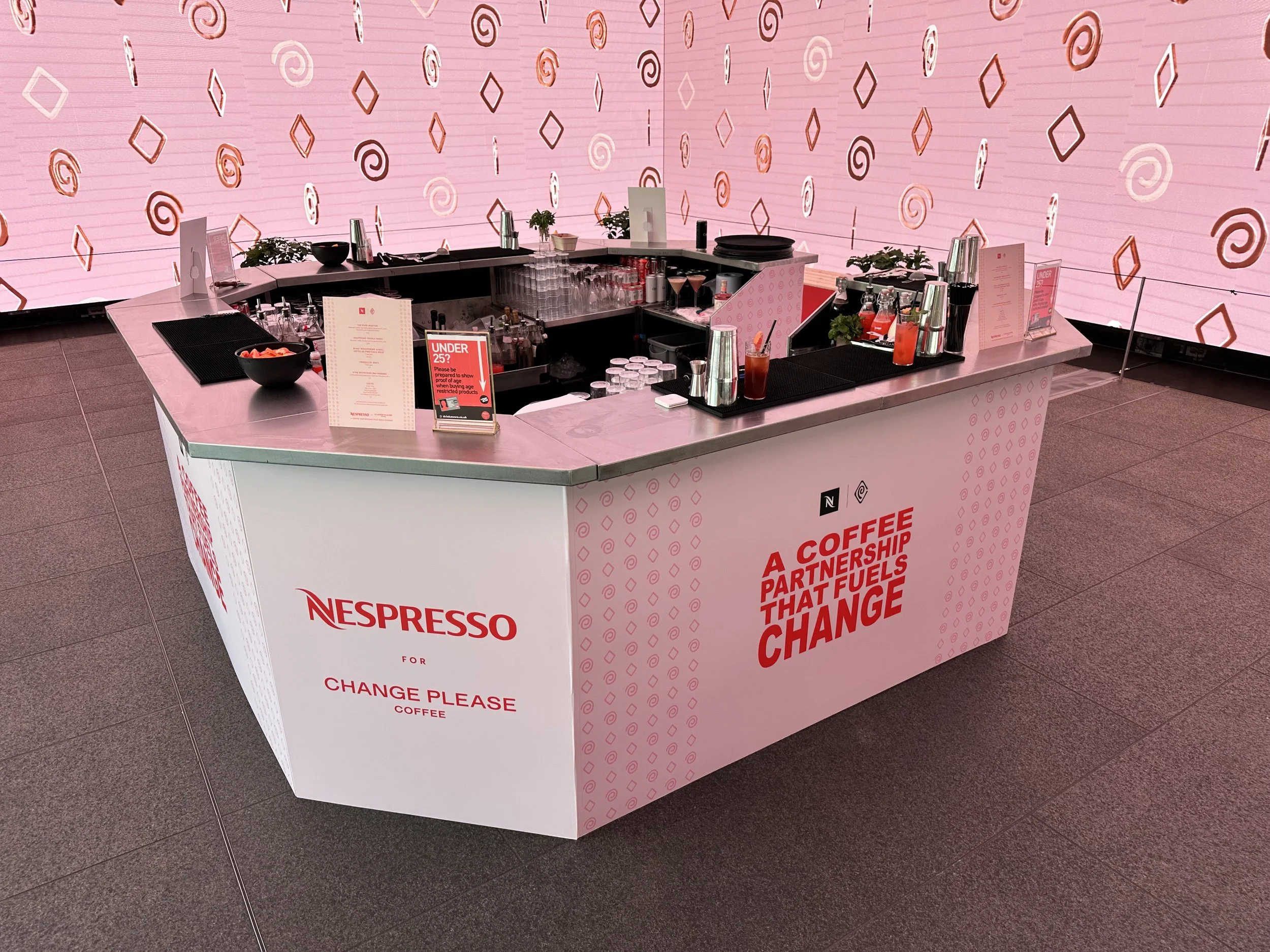A Nespresso coffee station with a pink and white color scheme, featuring a sign that says 'Nespresso for Change Please Coffee' and another with the message 'A coffee partnership that fuels change'. The station has various drinks, cups, and bar tools,