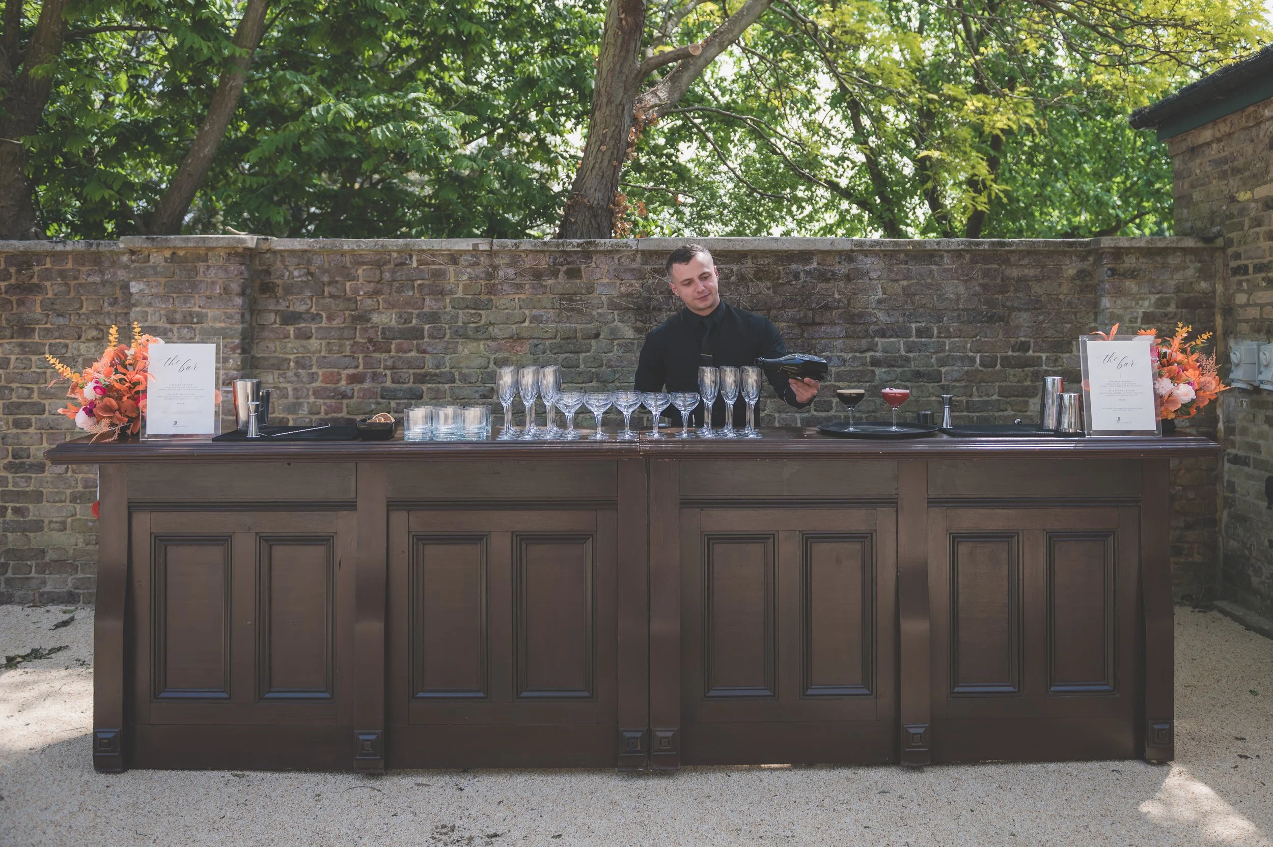 A bartender preparing drinks behind a wooden bar outdoors, with glassware and drink ingredients, flanked by floral arrangements on either side, against a brick wall and green trees.