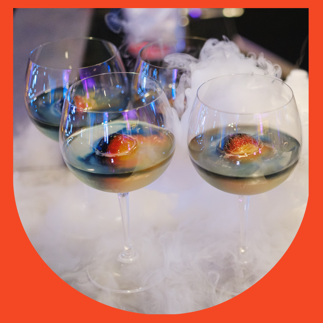 Four wine glasses filled with a clear liquid, each garnished with a strawberry and blueberry, with smoke or fog swirling around them, on a white surface.