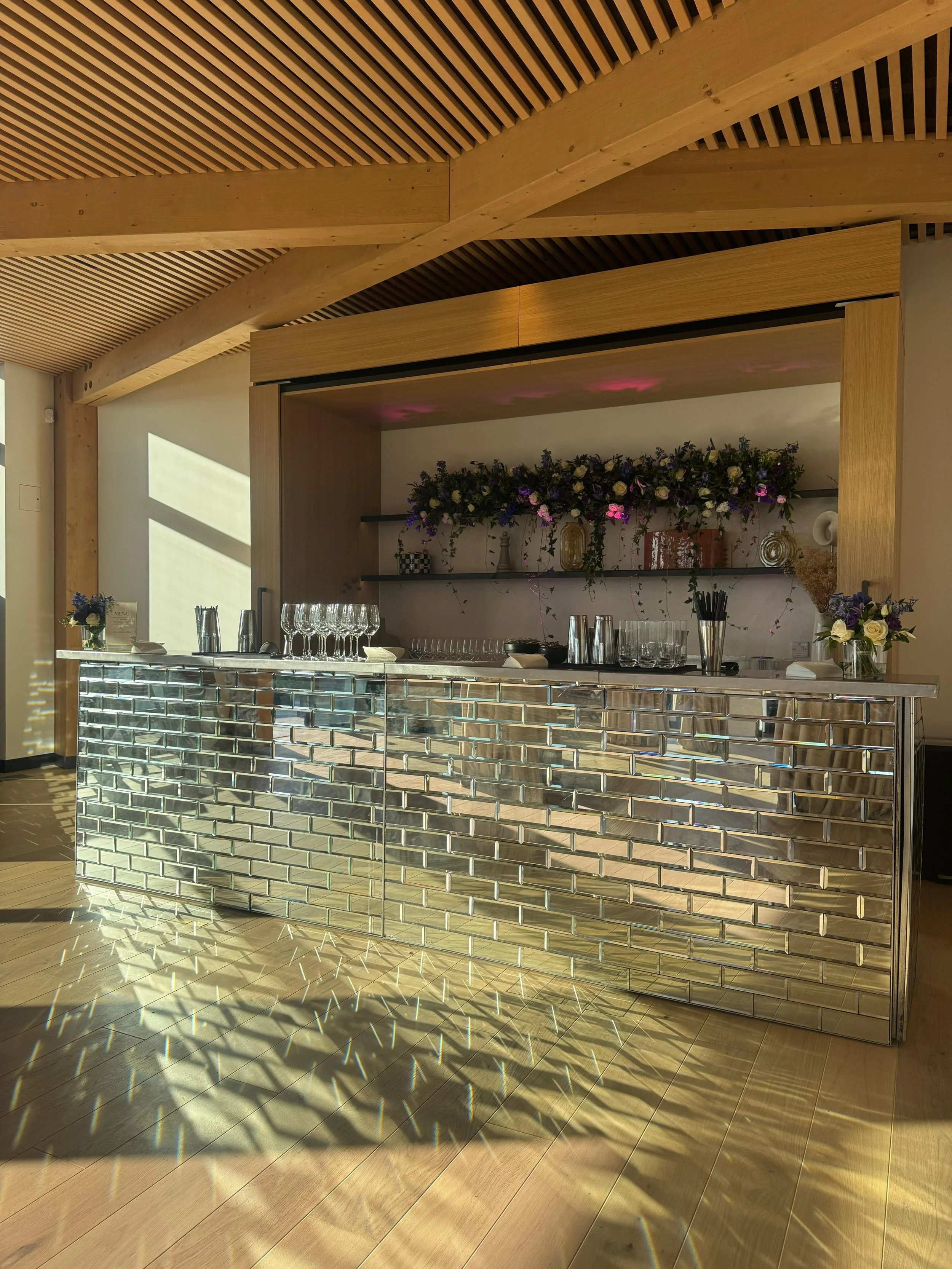 A modern bar with a metallic tiled front, set in a room with wooden ceiling beams and a decorated shelf behind it, featuring flowers and decorative objects.
