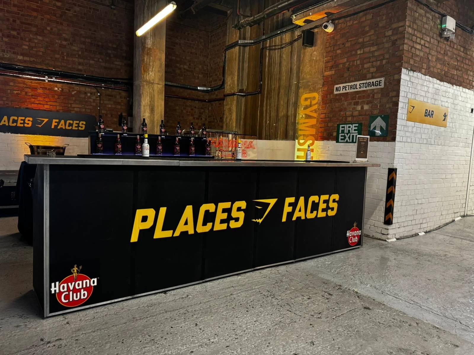 A bar or club scene with a black counter that has the words 'PLACES   FACES' in large yellow letters, with a Havana Club logo on the left and right corners. Behind the counter, there are several bottles lined up, a neon sign, and various neon and LED