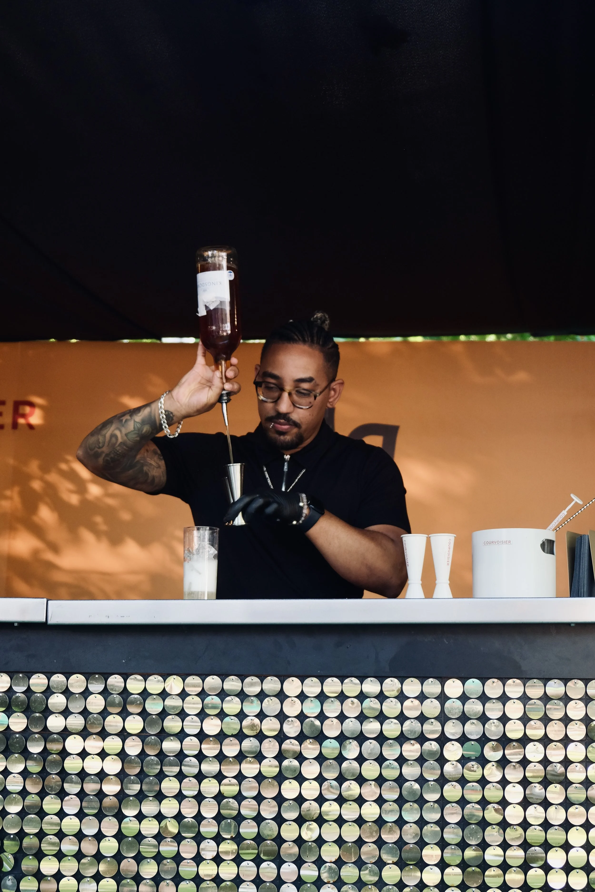 A bartender in black attire prepares a cocktail, pouring a dark liquid from a bottle into a jigger over a glass with a frothy drink at an outdoor bar.