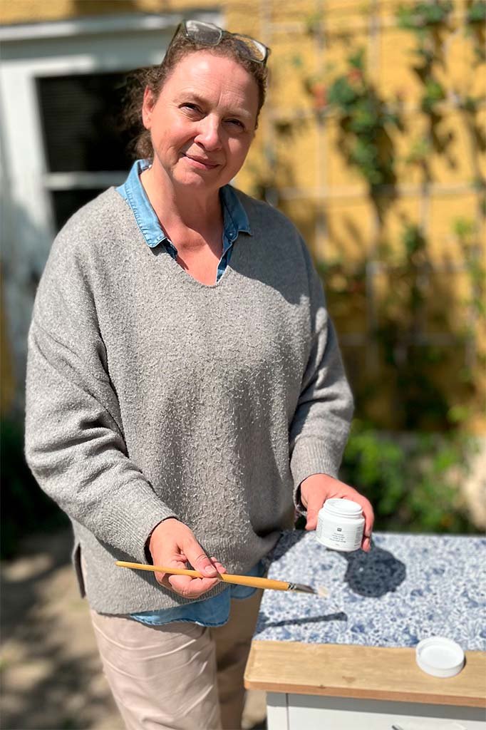 Frau steht im Garten mit Sonnenbrille auf dem Kopf, hält einen Pinsel und eine leere Dose, im Hintergrund ein gelbes Haus und grüne Pflanzen.