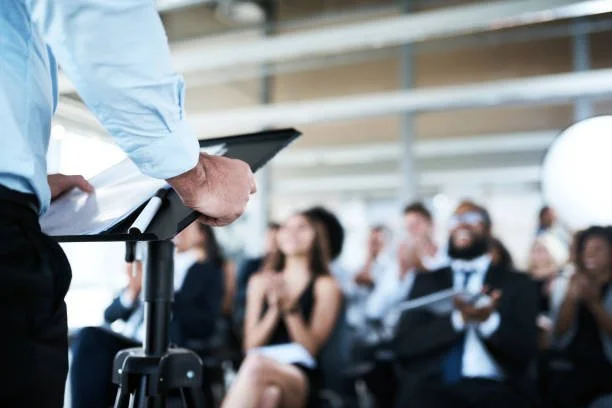 A person standing at a lectern giving a presentation to a seated audience in a conference room.