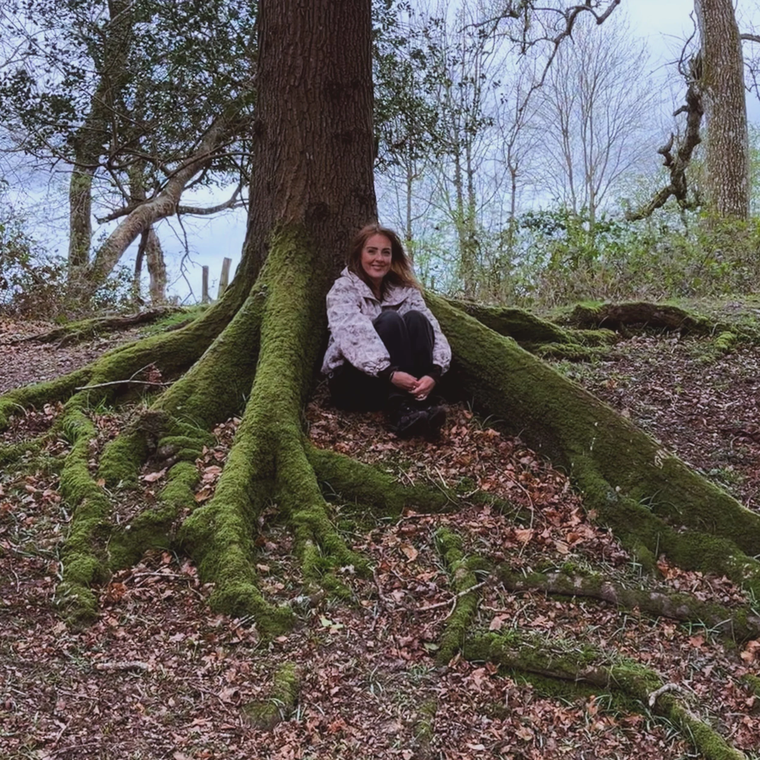 A woman sitting on the mossy roots of a large tree in a forest with bare trees in the background and a cloudy sky