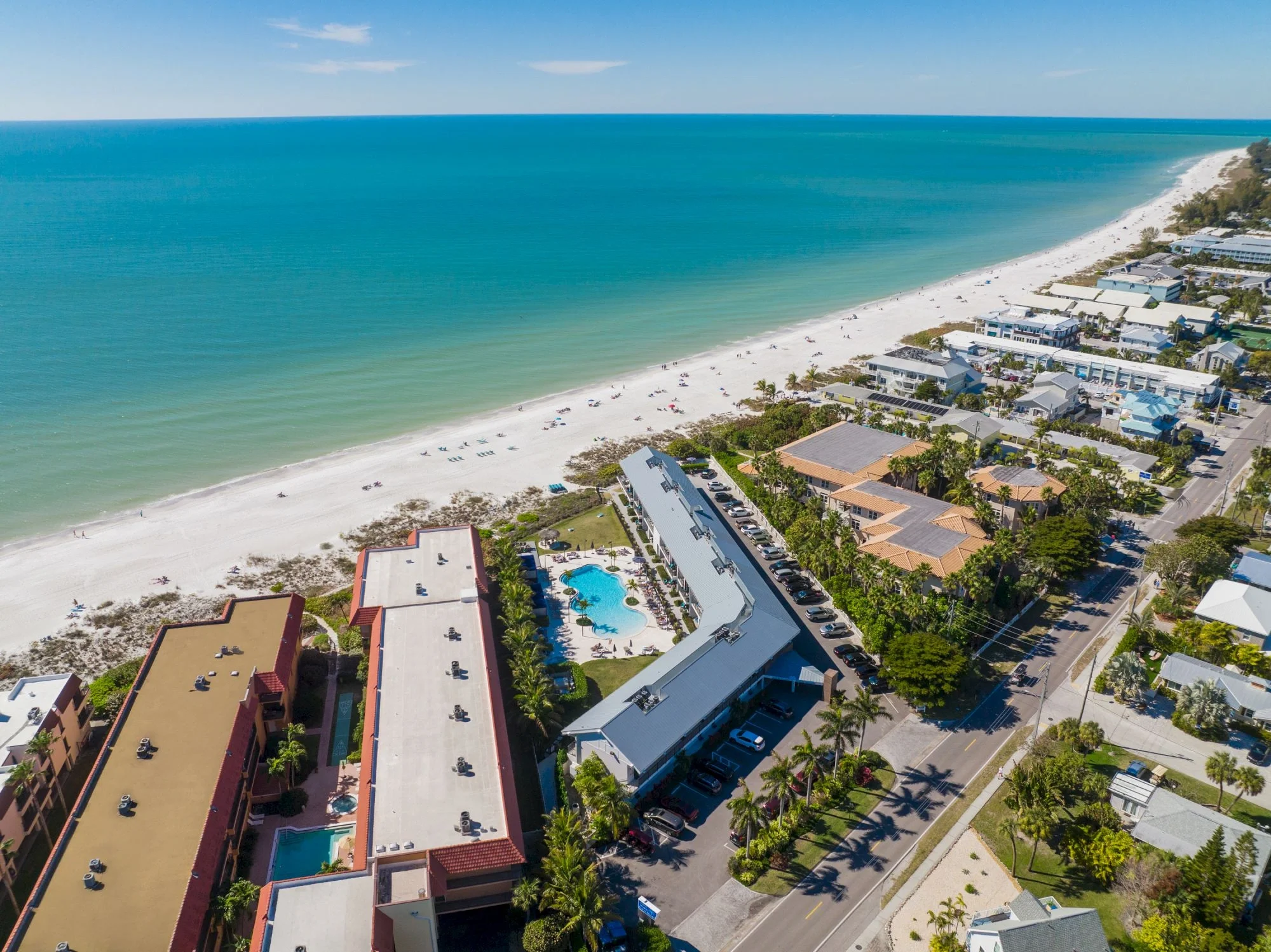 Sarasota Motor Club - Exotic Car Rental Sarasota Florida - Aerial view of a beachside resort with a large hotel, swimming pools, and a sandy beach with umbrellas and lounge chairs, facing the ocean with turquoise waters and a clear sky.