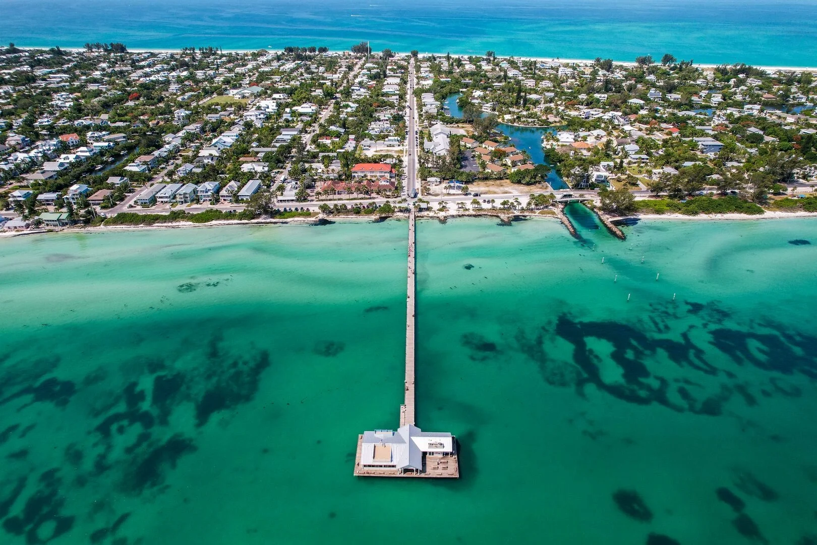 Aerial view of a coastal town with a long pier extending into turquoise water, leading to a white building on a small platform at the end of the pier. Residential houses and a canal system are visible behind the town, with the ocean in the background.