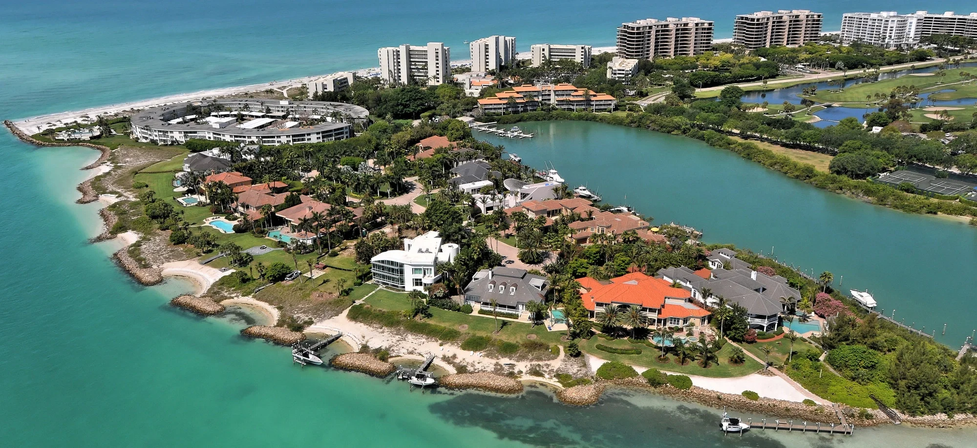 Sarasota Motor Club - Sports Car Rental Sarasota FL - Aerial view of a coastal residential area with houses, pools, and boats along the shoreline, with high-rise buildings and a golf course in the background.