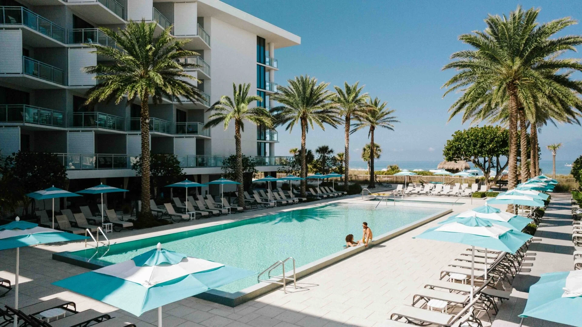 Sarasota Motor Club - Sports Car Rental Sarasota FL - A swimming pool area at a modern hotel with lounge chairs and blue umbrellas, surrounded by palm trees, with a view of the ocean in the background.