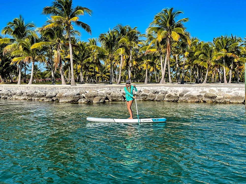 Sarasota Motor Club - Exotic Car Consignment Sarasota - A woman on a paddleboard in clear water near a rocky shore, with a background of palm trees and a bright blue sky.
