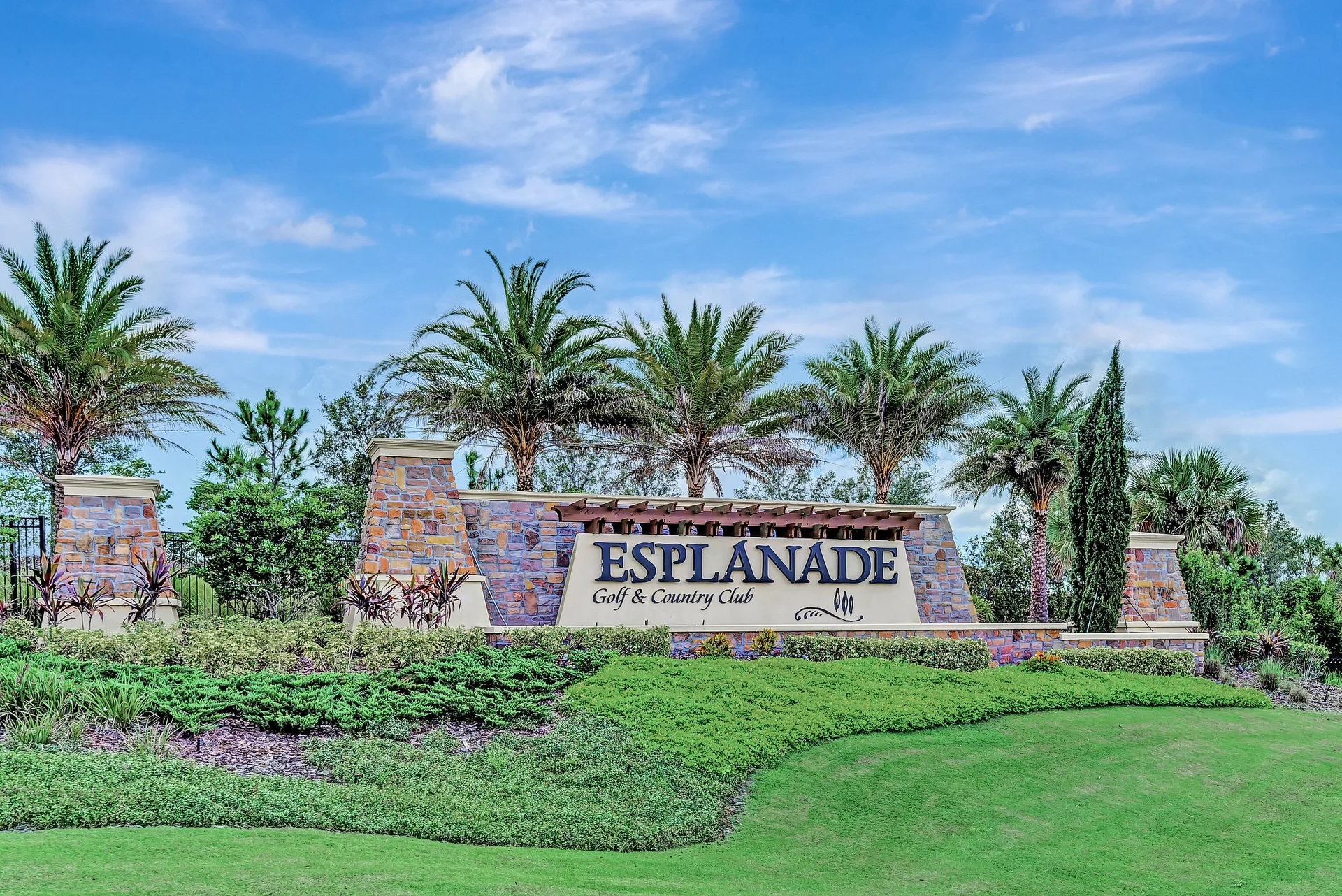 Sarasota Motor Club - Premium Car Service Sarasota - Main entrance sign for Esplanade Golf & Country Club with stone pillars, surrounded by green lawn, shrubs, and tall palm trees against a blue sky.