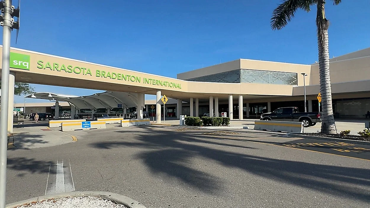 Sarasota Motor Club - Sports Car Rental Sarasota FL - Sarasota Bradenton International Airport entrance with a large green sign and covered drop-off area, a driveway, and a few cars parked nearby, under a clear blue sky with some palm trees.