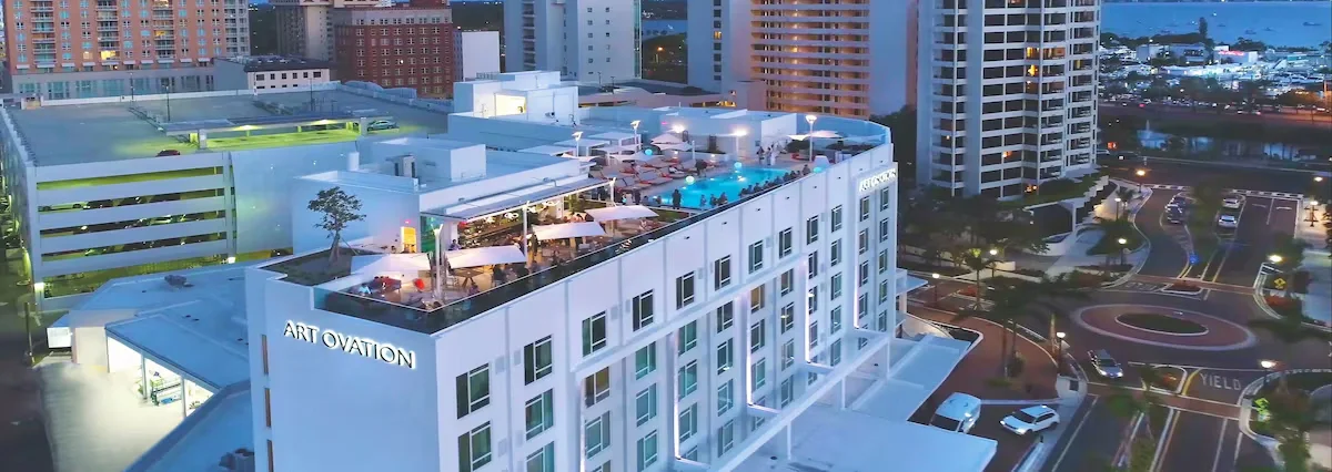 Sarasota Motor Club - premium car service sarasota - Nighttime aerial view of a white multi-story hotel named 'Art Ovation' with a rooftop pool area, umbrellas, and lounge chairs in a city with tall buildings and parking lot.