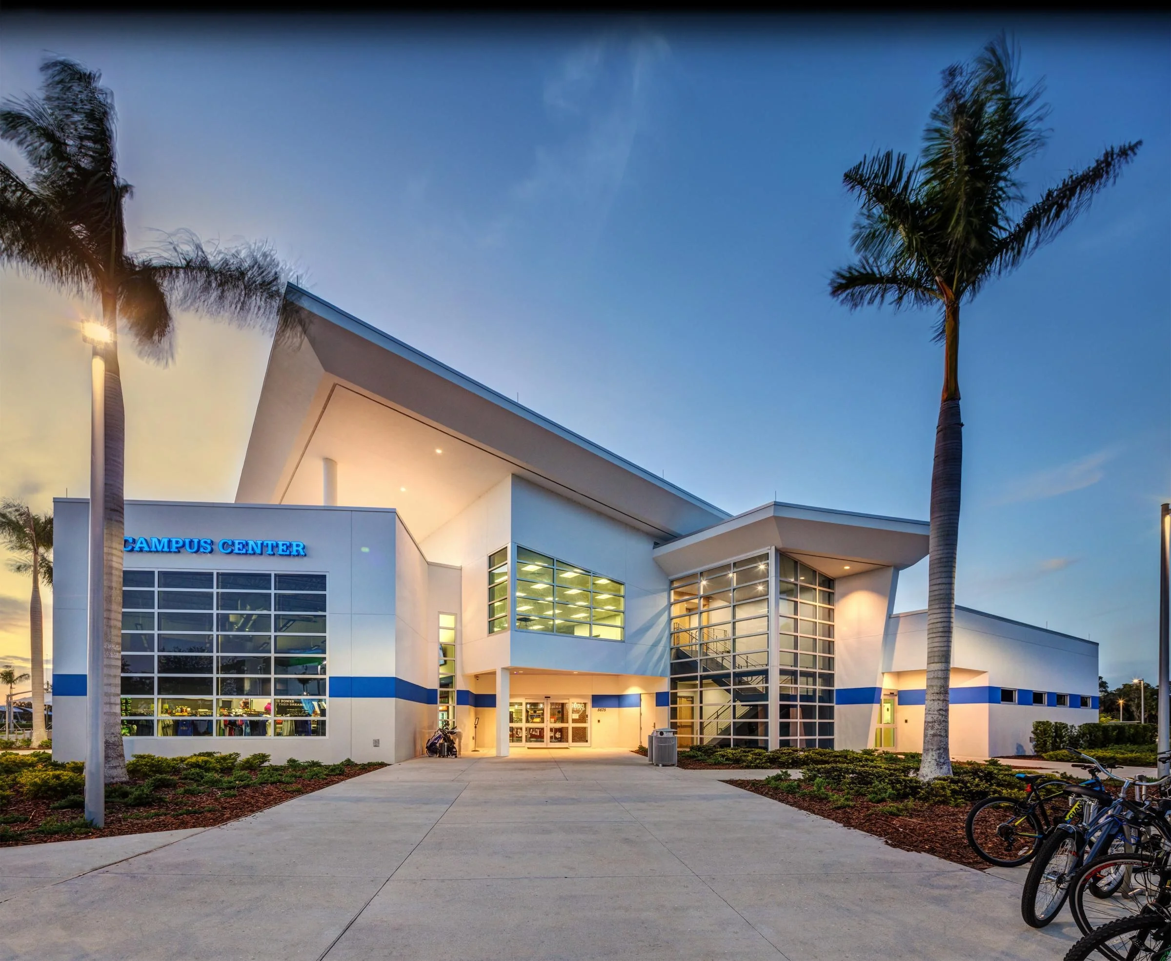 Sarasota Motor Club - Sports Car Rental Sarasota FL - Modern white campus center building with large glass windows, palm trees, and bicycles outside during evening.