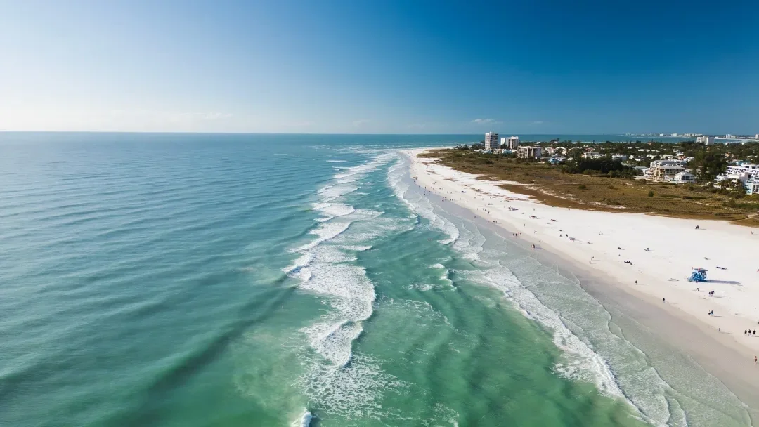 Sarasota Motor Club - Premium Car Service Sarasota - Aerial view of a beach with white sand, waves, and a line of buildings in the distance on a sunny day.