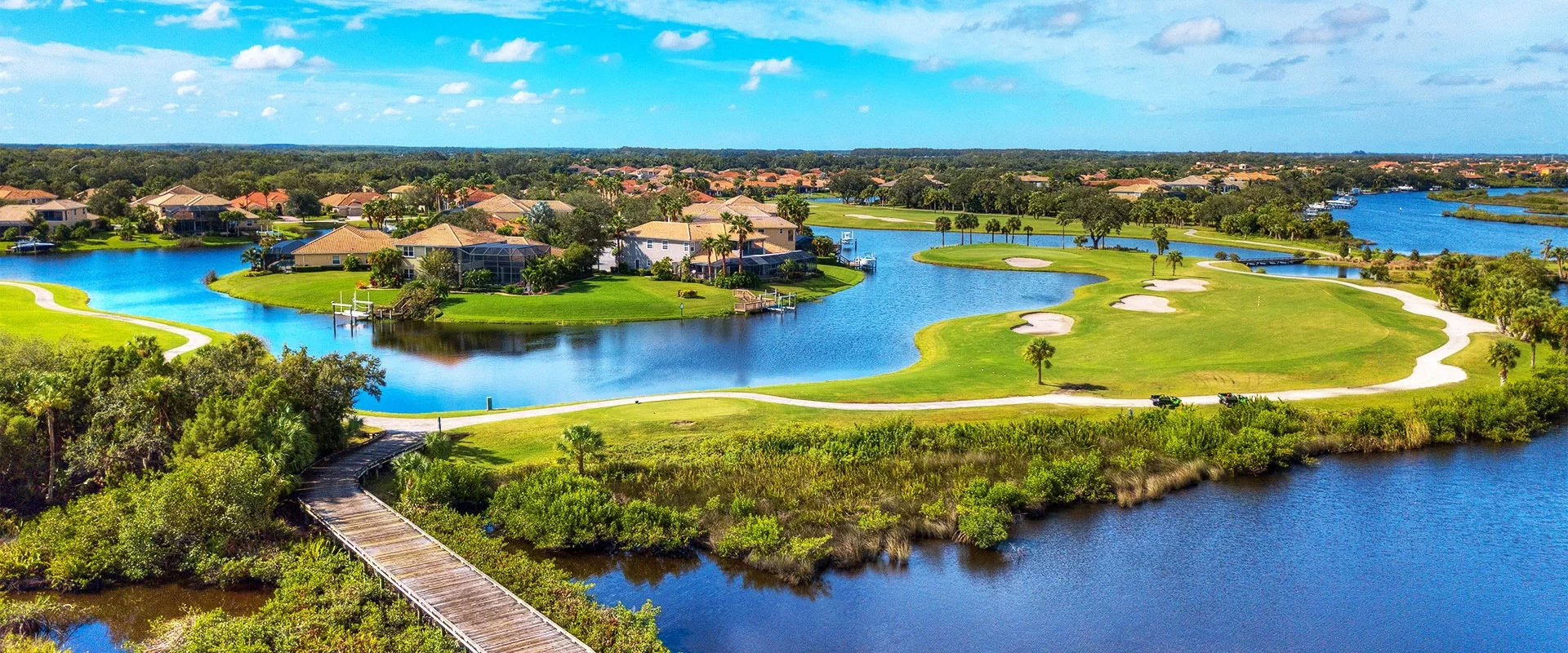 Sarasota Motor Club - Exotic Car Rental Sarasota FloridaA scenic aerial view of a residential golf course with water hazards, sandy bunkers, lush green fairways, houses with screened porches, and a wooden bridge over water, under a partly cloudy sky.