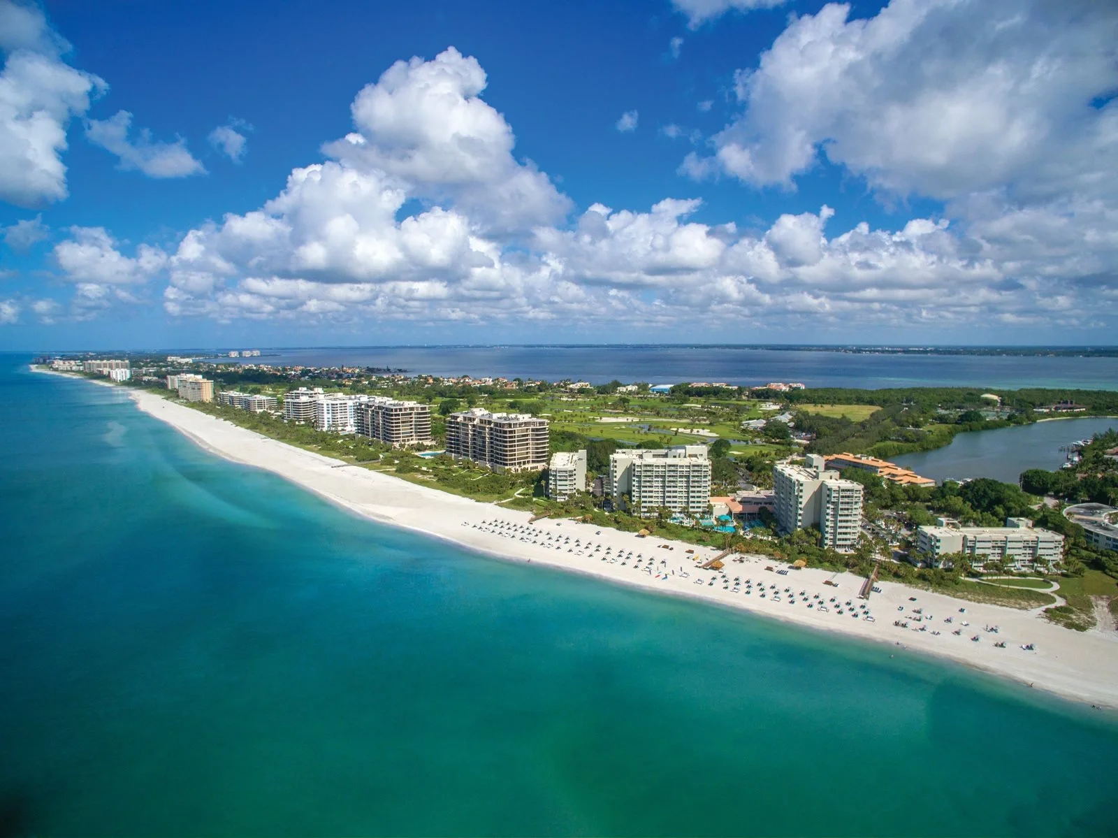 Sarasota Motor Club - Premium Car Service Sarasota - Aerial view of a beach with white sand, turquoise water, and beach umbrellas, lined with modern beachfront buildings, green parks, and a river in the background under a partly cloudy sky.