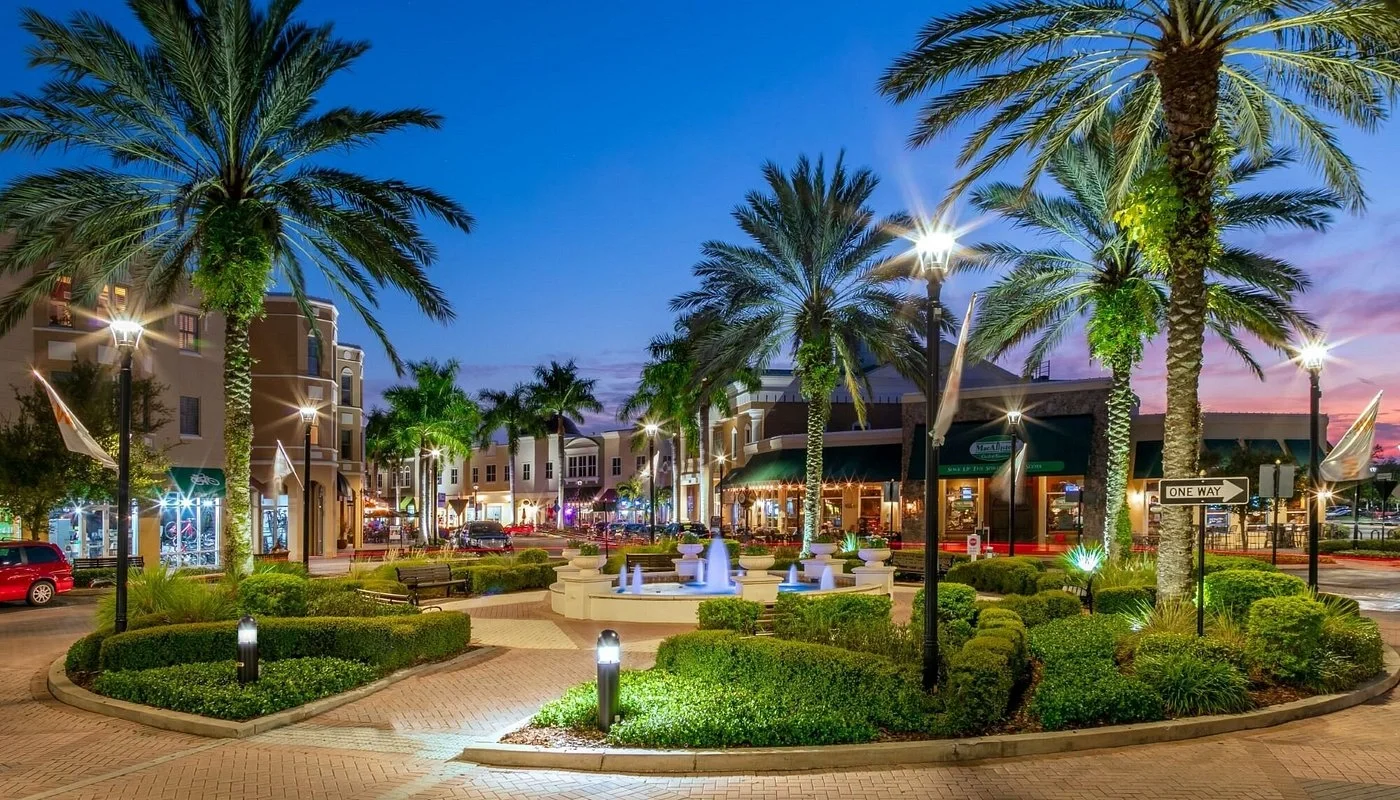 Outdoor shopping area at dusk with palm trees, a fountain, and lit walkways in a modern commercial district.