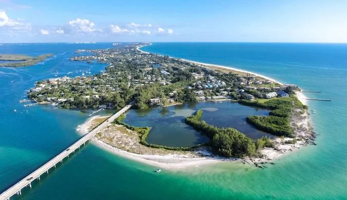 Sarasota Motor Club - Exotic Car Rental Sarasota Florida - Aerial view of a coastal island with a bridge connecting it to the mainland, featuring lakes, green areas, sandy beaches, and the ocean under a blue sky with some clouds.