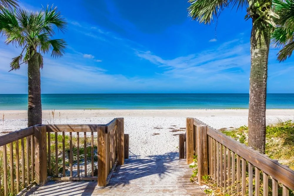 Sarasota Motor Club - Premium Car Service Sarasota - View from wooden steps onto a sandy beach with turquoise ocean and blue sky, flanked by palm trees.