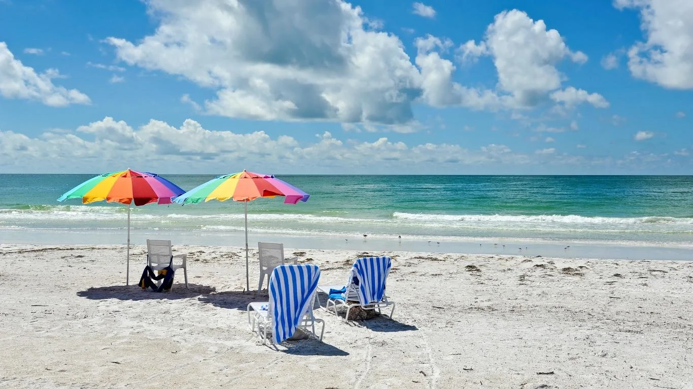 Sarasota Motor Club - Premium Car Service Sarasota - Empty beach scene with colorful umbrellas and chairs facing the ocean, with a partly cloudy sky.
