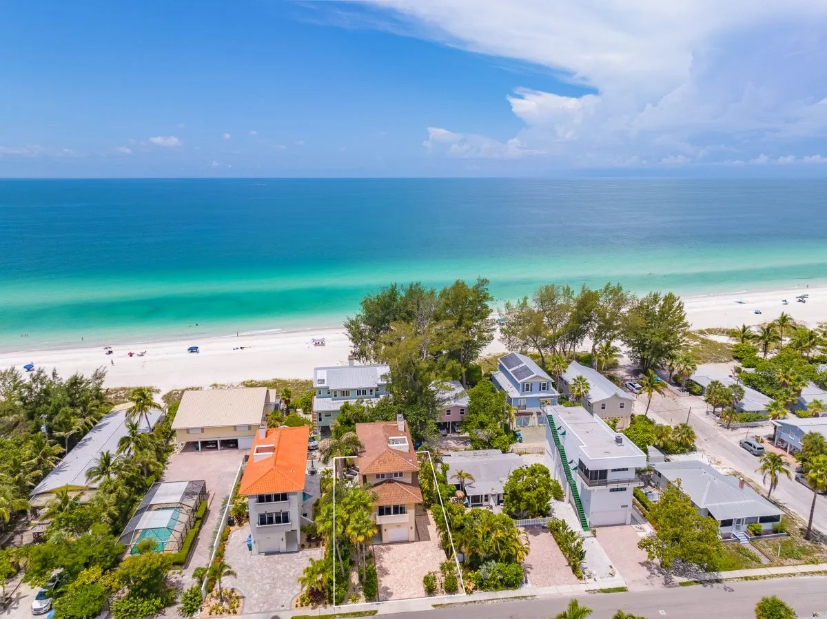 Sarasota Motor Club - Premium Car Service Sarasota - Aerial view of a beach with turquoise water, white sandy shore, and houses with trees and greenery along the coast.