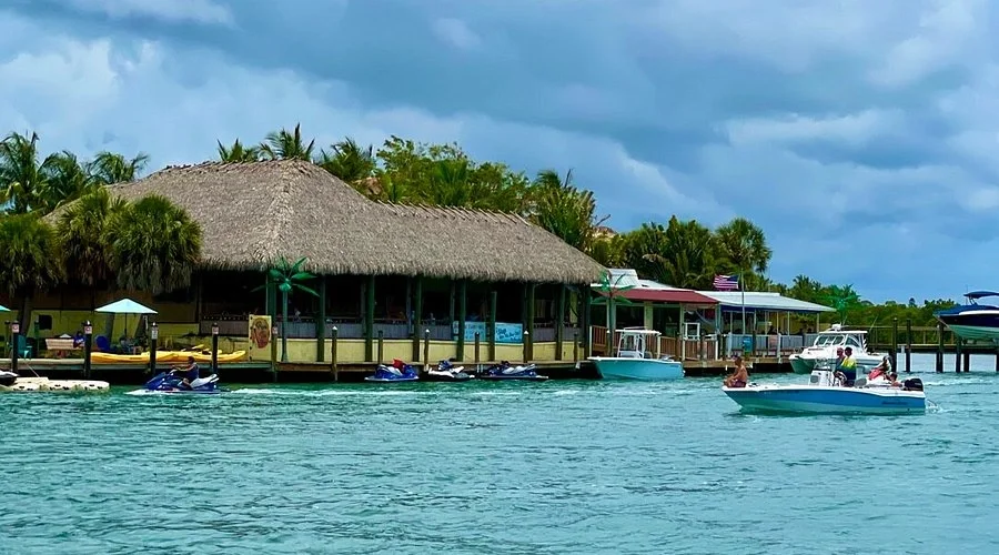 Sarasota Motor Club - Premium Car Service Sarasota - A tropical waterside scene with boats, jet skis, acrobatic watercraft, a thatched-roof restaurant, and lush palm trees under a cloudy sky.