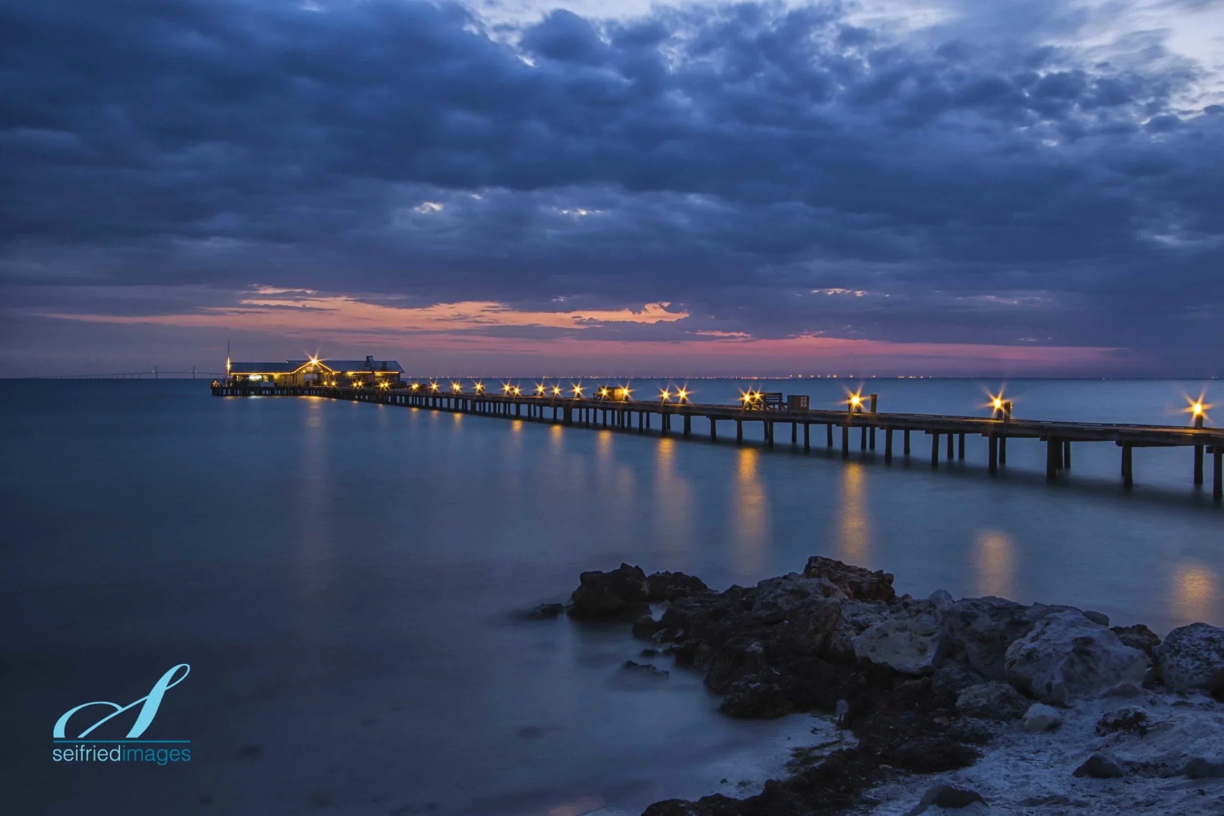 A long pier extends into a calm body of water during dusk, with a small building at the end illuminated by yellow lights. The sky is partly cloudy with shades of pink, purple, and dark blue, and reflections of the lights are visible on the water surface.