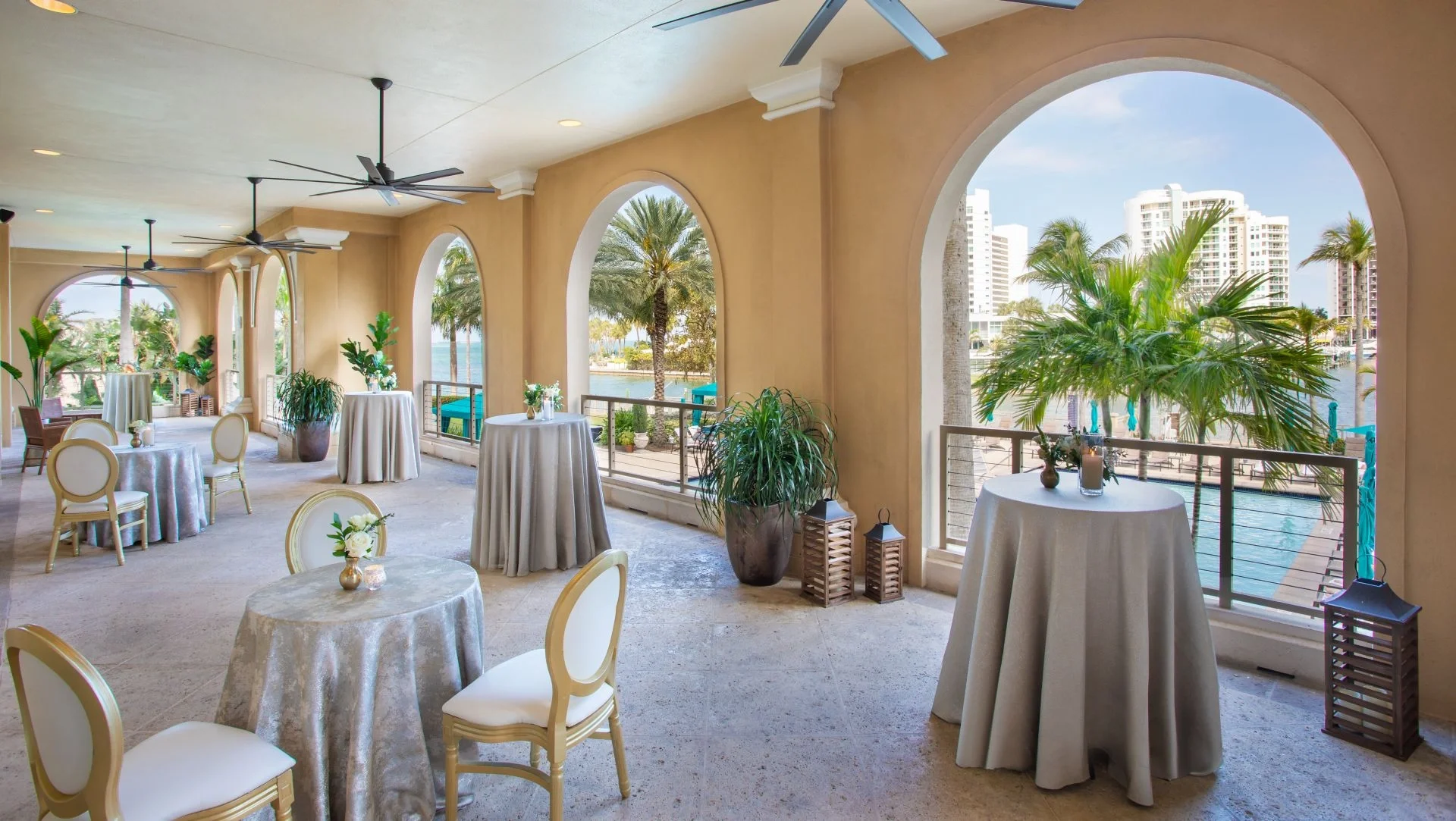 Elegant outdoor balcony with round tables covered in light-colored tablecloths, decorated with small floral centerpieces and candles, surrounded by white cushioned chairs, overlooking a waterfront with palm trees and high-rise buildings in the background.