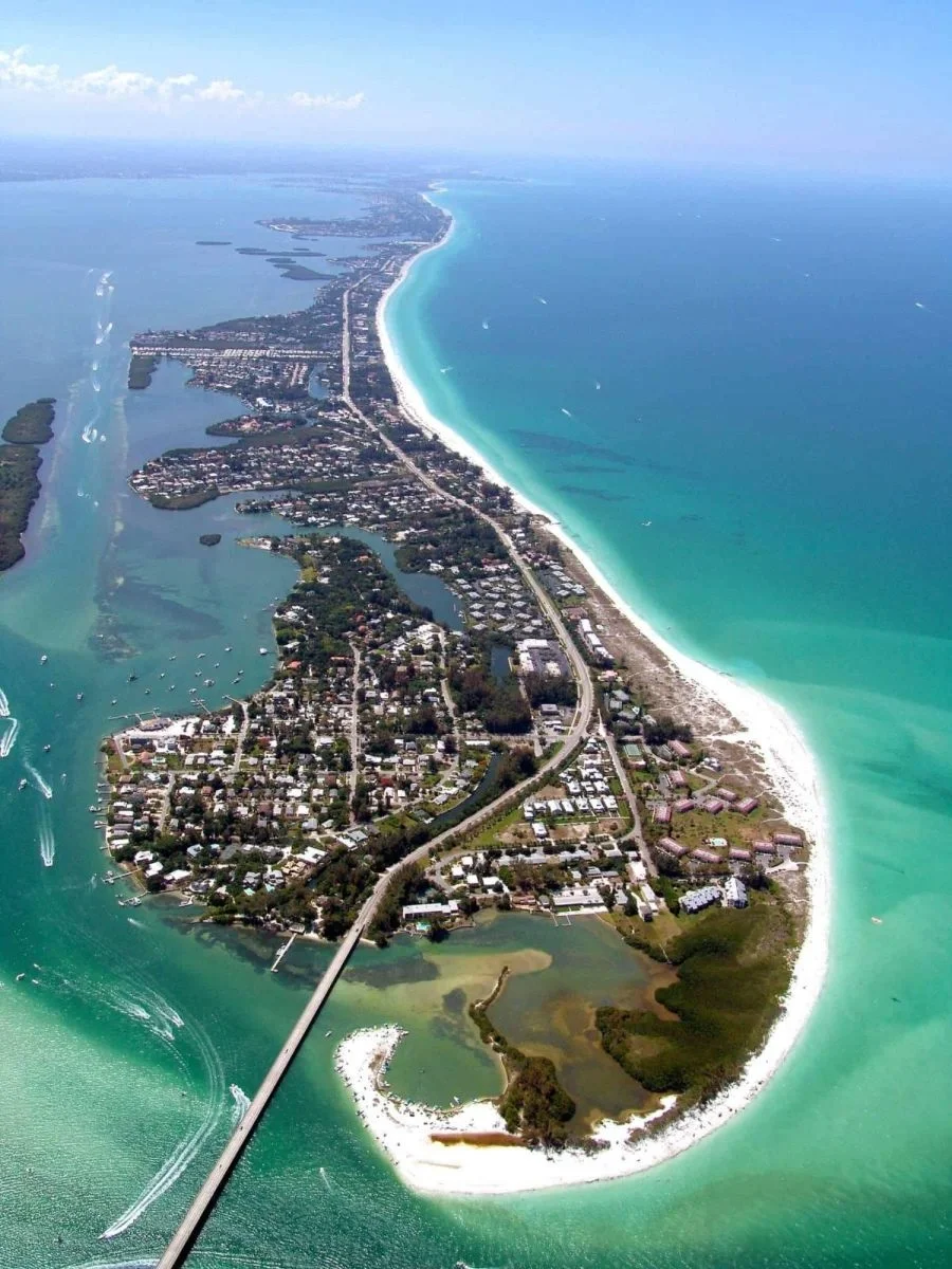 Sarasota Motor Club - Exotic Car Consignment Sarasota - Aerial view of a coastal area with a long sandy beach, a bridge connecting landmasses, residential neighborhoods, and turquoise ocean waters.