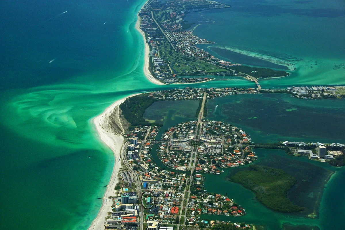 Sarasota Motor Club - Exotic Car Consignment Sarasota - Aerial view of a coastal city with turquoise waters, white sandy beaches, and urban area along the shoreline and inland, showing water outlets and green spaces.