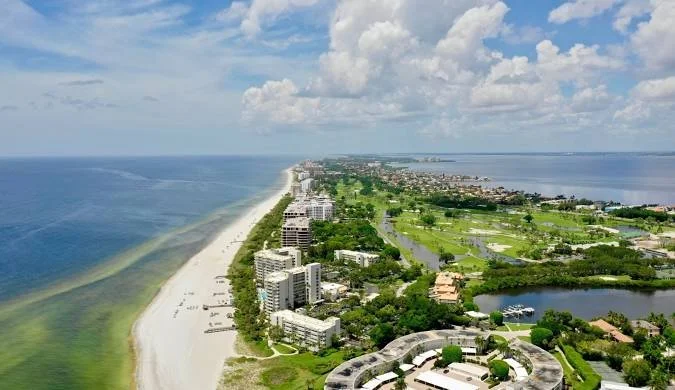 Sarasota Motor Club - Exotic Car Rental Sarasota Florida - Aerial view of a coastal city with high-rise buildings, a sandy beach, and lush greenery, with a large body of water on the left and a river or lagoon on the right, under a partly cloudy sky.