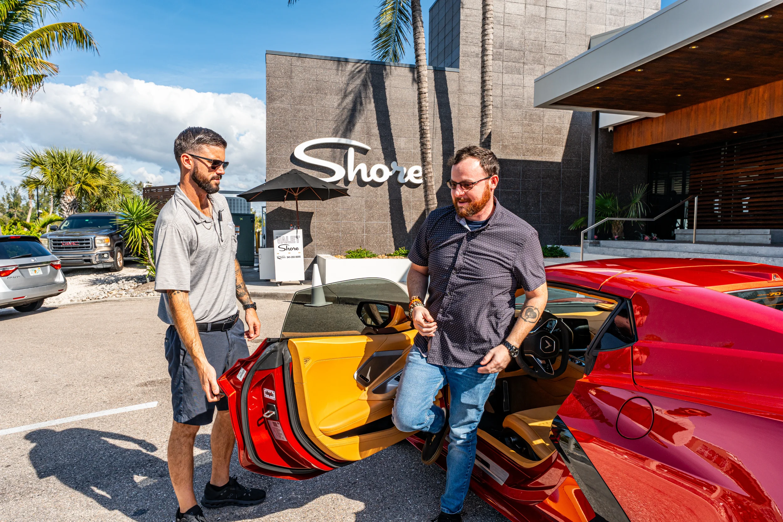 Sarasota Motor Club - exotic car rental - Two men standing next to a red Corvette with a yellow interior, outside a building with a sign that says "Shore". One man is opening the car door and the other is looking inside.