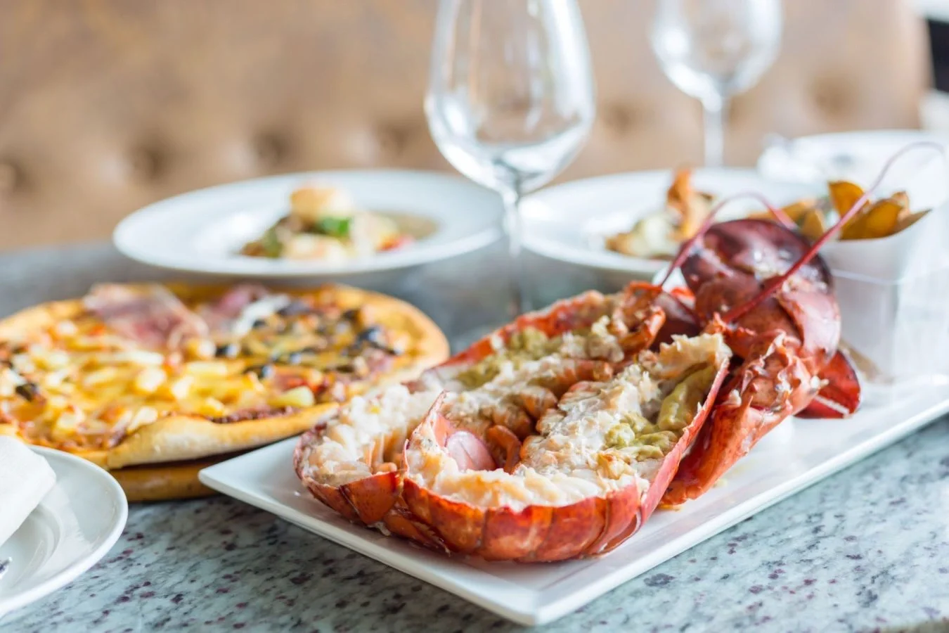Sarasota Motor Club - Exotic Car Consignment Sarasota - Lobster tail on a white platter with butter, alongside a pizza with cheese, ham, and vegetables, on a marble table in a restaurant setting.