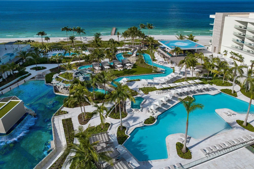 Sarasota Motor Club - Exotic Car Rental Sarasota Florida - Aerial view of a luxury beachfront hotel with multiple pools, palm trees, and a sandy beach and ocean in the background.