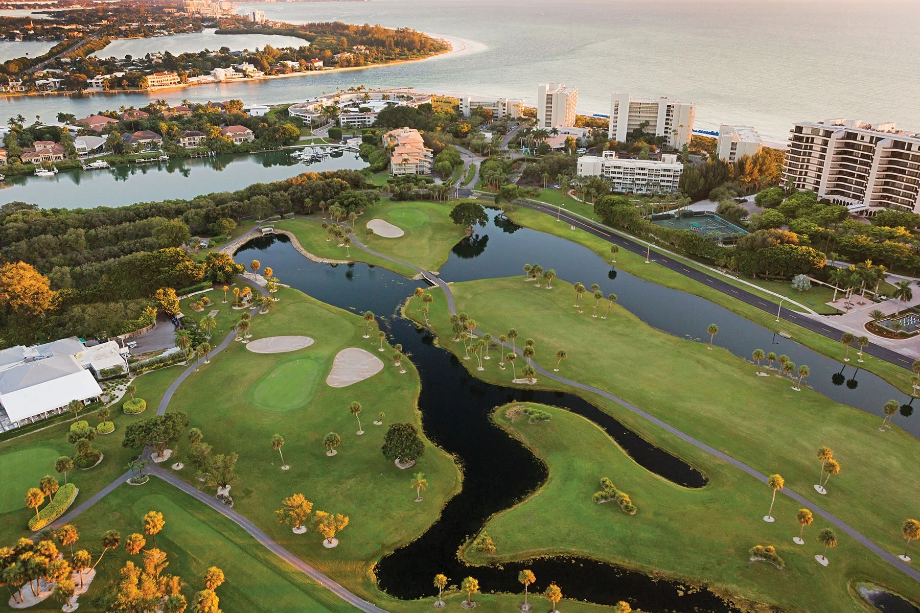 Sarasota Motor Club - Sports Car Rental Sarasota FL - Aerial view of a golf course with multiple water hazards, surrounded by trees, residential buildings, and a coastline with the ocean in the background.