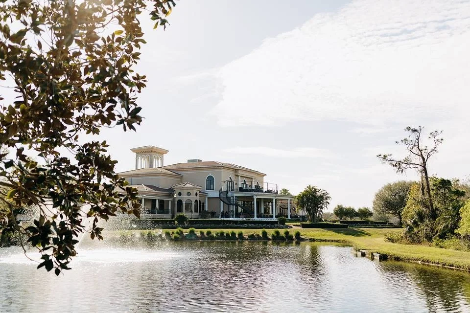 Sarasota Motor Club - Premium Car Service Sarasota - Large luxurious mansion with a prominent tower, situated by a reflective pond with fountain, surrounded by green lawn and trees.