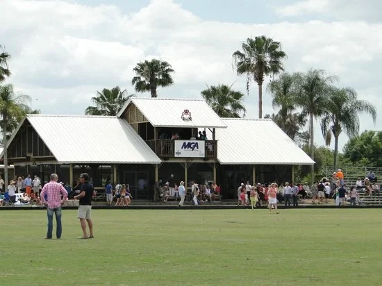 Sarasota Motor Club - Exotic Car Rental Sarasota Florida - A crowd gathered outside a large wooden clubhouse with a white metal roof, surrounded by palm trees, on a cloudy day at a golf course.