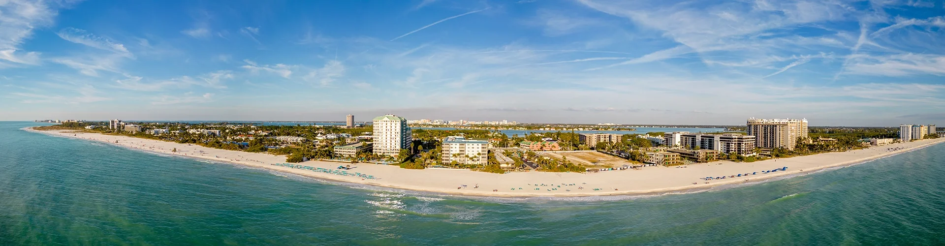 Sarasota Motor Club - Exotic Car Consignment Sarasota - A panoramic view of a tropical beach with white sand, turquoise water, and high-rise buildings in the background under a partly cloudy sky.
