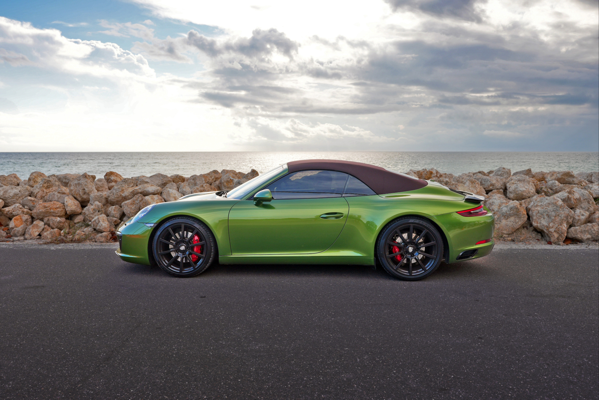 Sarasota Motor Club - Premium Car Service Sarasota - Green convertible sports car parked on a paved road near rocky shoreline overlooking ocean with cloudy sky.