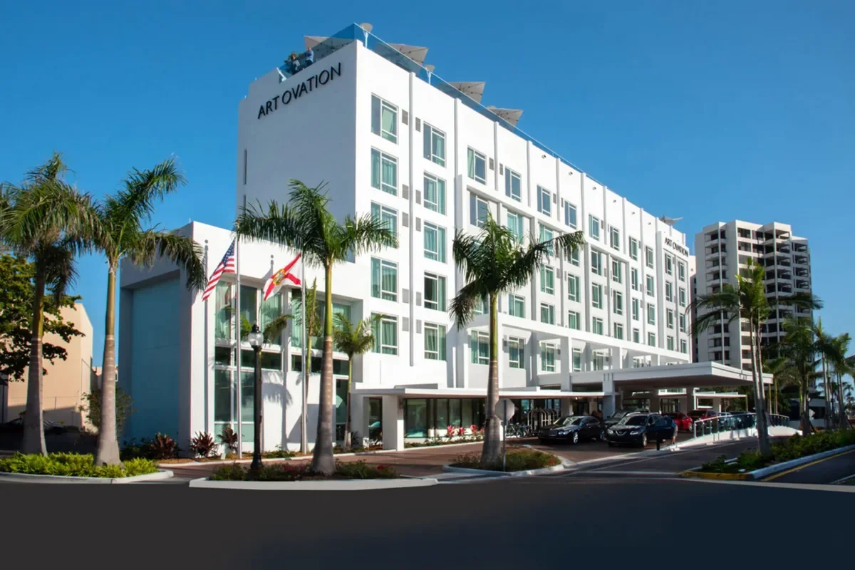 Sarasota Motor Club - Sports Car Rental Sarasota FL - Modern white multi-story hotel building with palm trees in front and parked cars, labeled ART OVATION, under a blue sky.