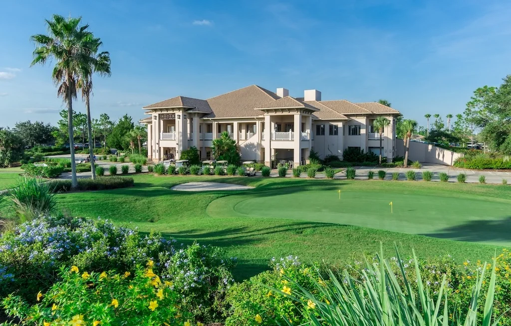 Sarasota Motor Club - Premium Car Service Sarasota - Large house with multiple balconies overlooking a golf course with a putting green, palm trees, and landscaped garden with flowering plants.