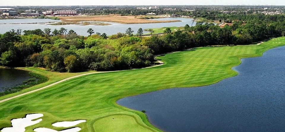 Sarasota Motor Club - Exotic Car Rental Sarasota Florida - A scenic golf course with lush green fairways, water hazards, and surrounding trees, with a distant cityscape and a body of water in the background.
