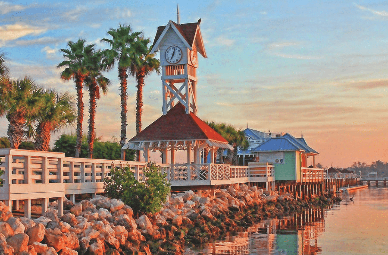 Sarasota Motor Club - Premium Car Service Sarasota - Sunset view of a waterfront with a white wooden pier, a small clock tower with a red roof, palm trees, and a colorful building in the background, reflecting on the water.