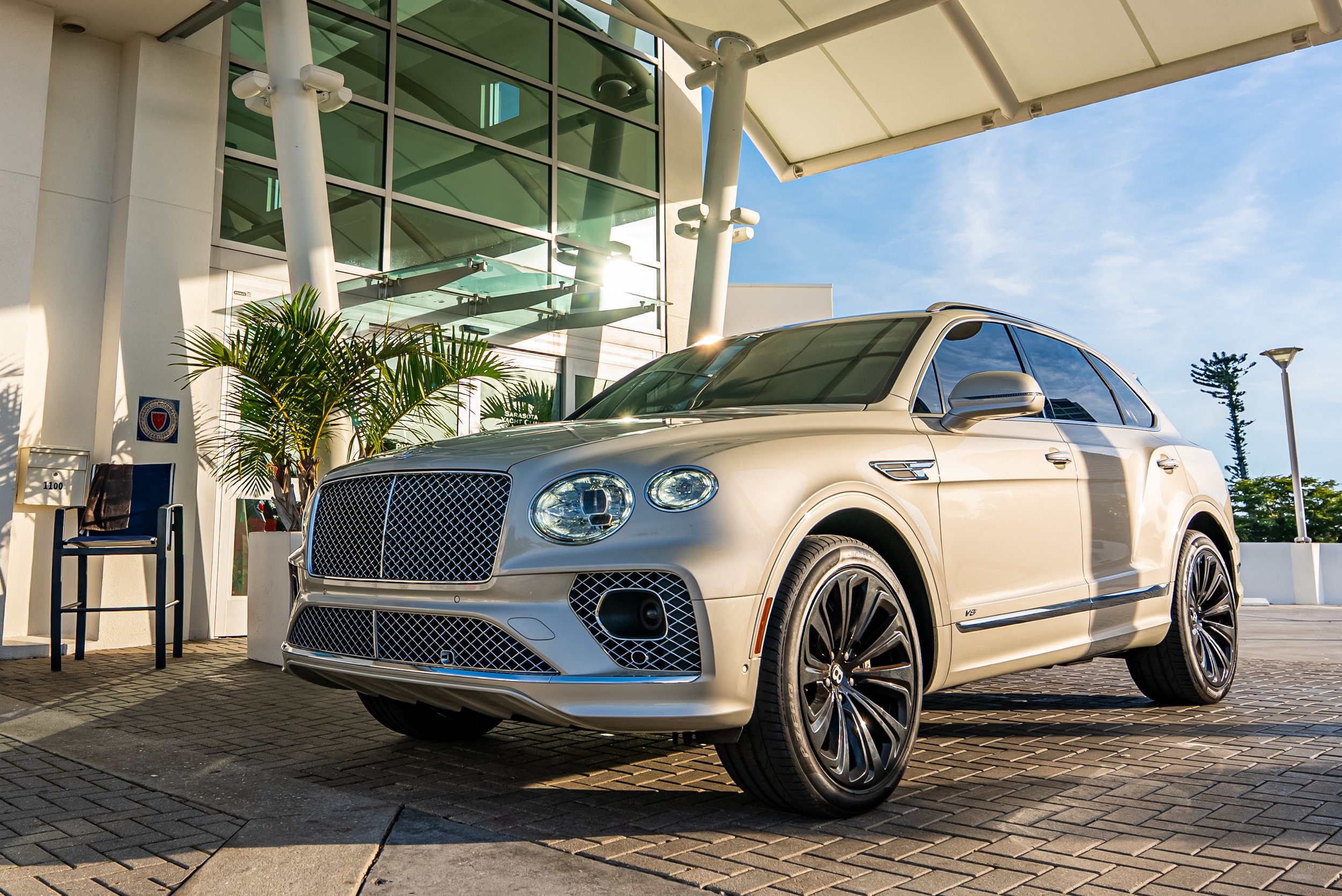 Sarasota Motor Club -  luxury car rental sarasota - Luxury silver Bentley Bentayga parked outside a modern building with glass walls, a seating chair, and potted plants, under a clear sky.