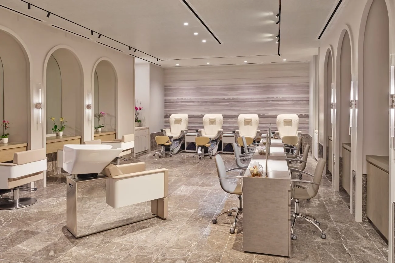 Sarasota Motor Club - Exotic Car Rental Sarasota Florida - Interior of a modern nail salon with pedicure chairs in the background, manicure stations with chairs in the foreground, and decorative plants along the walls.