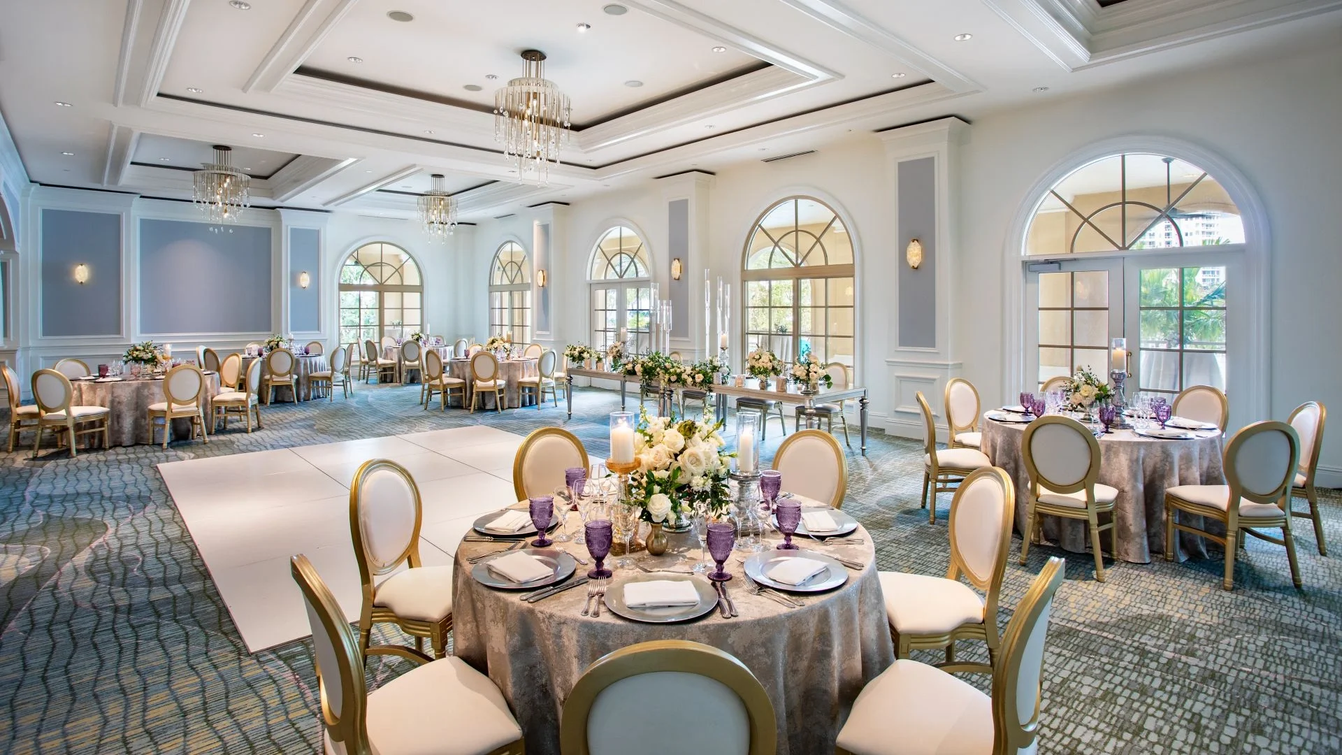 Elegant banquet hall decorated for a formal event with round tables, floral centerpieces, candles, and purple glassware, large arched windows, chandeliers, and white walls with molding.