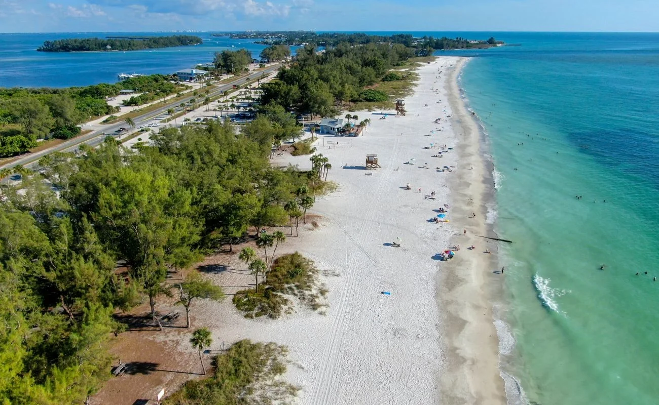 Sarasota Motor Club - Premium Car Service Sarasota - Aerial view of a sunny beach with white sand, scattered umbrellas and lounge chairs, surrounded by green trees and a calm blue ocean with people swimming and wading.
