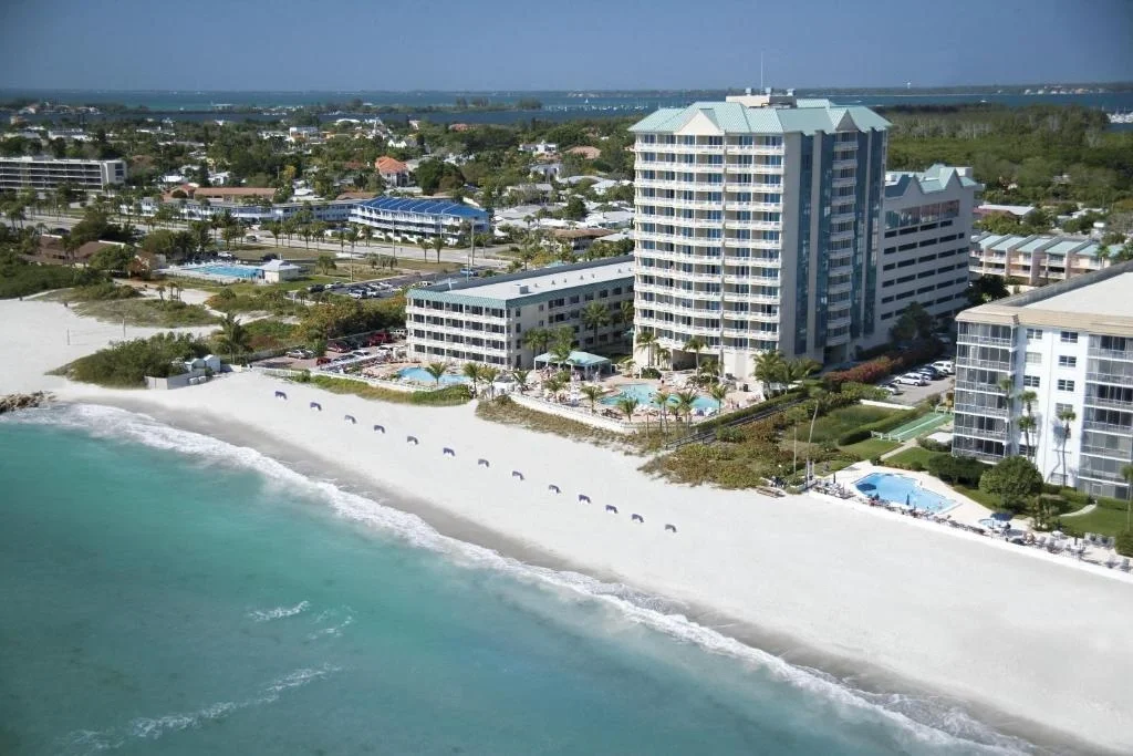 Sarasota Motor Club - Lido Beach Resort exotic car rental - Aerial view of a luxury beachfront resort with multiple swimming pools, palm trees, lounge chairs, and direct access to a sandy beach and blue ocean.